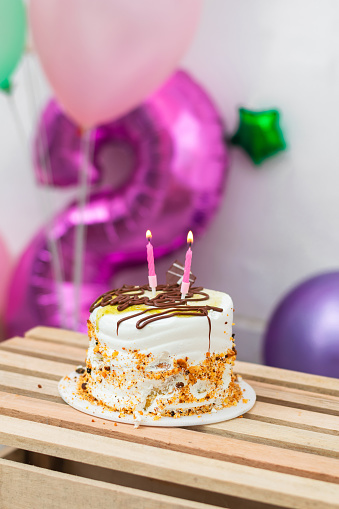 small birthday cake with two lit candles on it, on top of a wooden table with balloons in the background