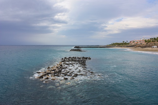 A stone jetty juts into the deep blue ocean with a sandy beach with hotels in the background. The sky is cloudy and the water is calm with small waves splashing on the rocks of the jetty.