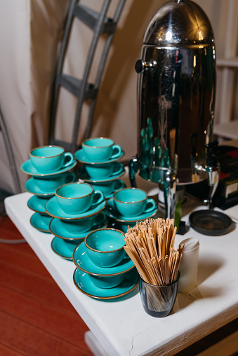 A coffee serving station featuring shiny metal coffee urn, turquoise ceramic cups with saucers, and wooden stir sticks, arranged neatly for a gathering.