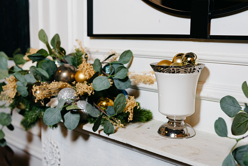 A classic holiday mantelpiece adorned with lush greenery, sparkling ornaments, and golden accents in an ornate white vase.