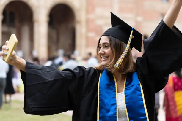 A young brunette adult taking a selfie with her hand up in the air in front of Royce Hall, UCLA on graduation day.