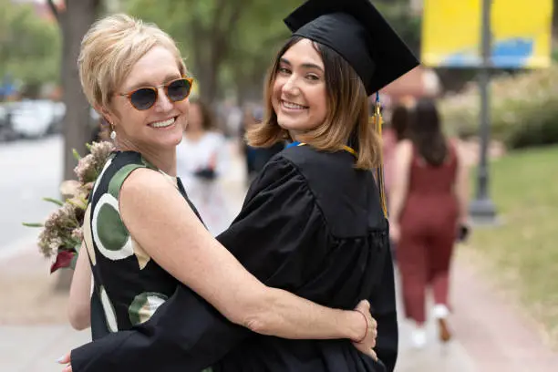 A brunette young adult in a graduation gown smiling while embracing with her mom on graduation day.