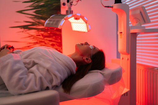 Side view of female patient undergoes red LED light or RLT therapy for skin rejuvenation at modern luxury aesthetic clinic. Young brunette woman client having non-invasive type of phototherapy.