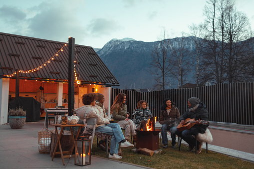 A family gathers around a firepit in their backyard during twilight, enjoying each other's company with the majestic mountains in the background, creating a cozy and memorable outdoor moment.