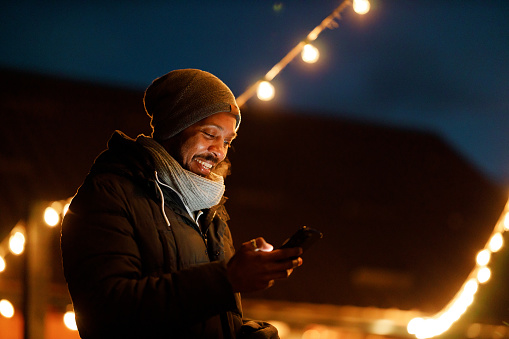 A black man, bundled up in warm winter attire, is captured enjoying a pleasant moment on his smartphone. His face is illuminated by the gentle glow of the screen and the festive lights overhead, contrasting with the darkening sky of a winter's evening.