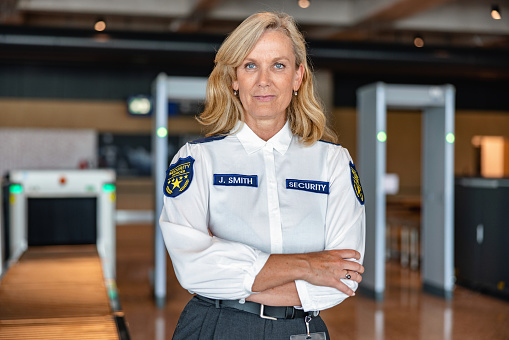 Portrait of a beautiful mid adult Caucasian female security worker with hands crossed posing for a photo in an airport lobby, looking at the camera, wearing a uniform shirt with name tag. Waist up shot.