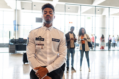 Portrait of a mid adult Black male security worker posing for a photo in an airport lobby, looking at the camera, wearing a uniform shirt and an ear piece. Waist up shot.