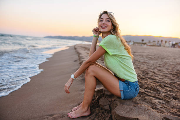 Happy Caucasian female tourists sitting on a beach in Barcelona and admiring the beautiful view of the sunset. She is sitting on the sand in her clothes and smiling. The woman looks happy and relaxed.