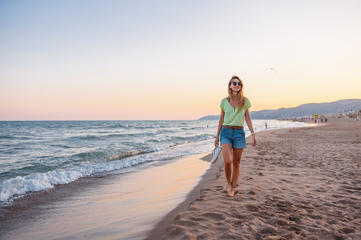 Attractive young adult Caucasian female enjoying her walk on the sandy beach. The sun is setting and creating a relaxing and calm atmosphere. The woman is walking barefoot while holding her shoes in one hand. She is spending her time alone at the beach. The woman is dressed in casual summer clothes.