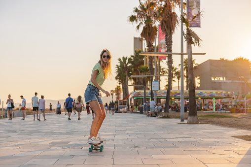 Sporty young adult Caucasian female having fun while riding a mini skateboard by the Barceloneta beach. She is located on a boardwalk surrounded by high palm trees. The sun is setting creating a beautiful warm light. Fun summer activities for sporty people.