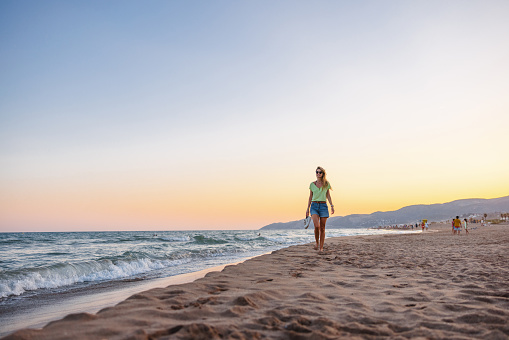 An attractive young adult Caucasian female enjoying her relaxing walk on the beach. She is walking barefoot while carrying her shoes in one hand. The sky is colorful due to the sunset. Fun summer activities to distress.
