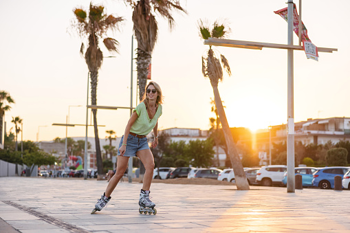 Attractive young adult female roller skater enjoying her evening at the beach. She is roller skating alone on the beach promenade at sunset. As the sun is setting it is creating a beautiful and relaxing golden light. The woman looks relaxed and content while rollerblading.