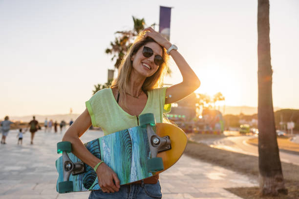 A portrait of a cheerful young adult Caucasian female skateboarder holding a cool and colorful penny board while smiling at the camera. She is located on the boardwalk by the beach. The sun is setting creating a relaxing atmosphere and a warm golden light.