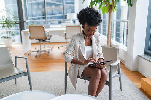 Serious and Focused Young Black Businesswoman working in her fancy office. The woman is sitting on the chair and using a digital tablet to read and respond to her e-mails. She is wearing an elegant business suit. The woman looks professional. Empowered black female CEO working on developing her business further.