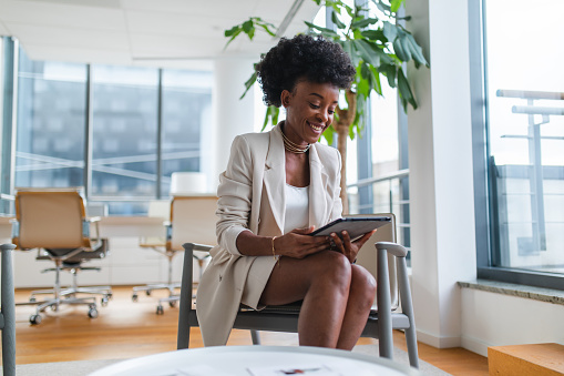 Happy black female businesswoman sitting on a chair in a modern office and looking at the tablet screen. She is smiling while going over this year's financial results. Successful female black manager doing a good job leading the company.