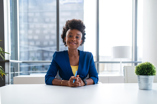 Successful black female manager looking at the camera and smiling while sitting by her desk at the office. She is wearing a fancy blue suit and looks elegant. Her office has a beautiful view of the city.