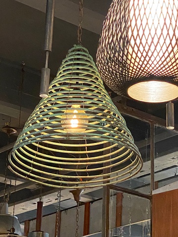 Looking up at two lanterns  hanging from ceiling in industrial building, one is a green conical spiral and the other is woven brown cylindrical shaped.