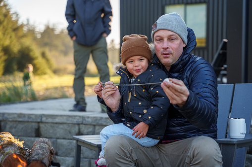An adult man teaches his toddler niece how to roast marshmallows while making s'mores in a campfire outside their rental house in the Pacific Northwest.