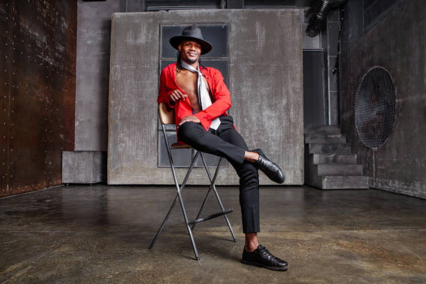 Contemporary African American man, actor or dancer sits on a stool on stage, dressed in a red shirt and a hat.