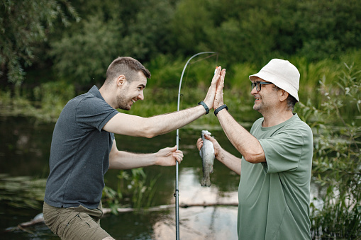 Father and adult son with fishing rods near the lake fishing at summer. Fishermen cath a fish and holding it. Men wearing khaki clothes and a caps.