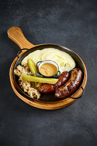 Top view of rustic grilled sausages with creamy mashed potatoes and tangy pickles, on a wooden server against a textured backdrop. Comfort cuisine at its finest.