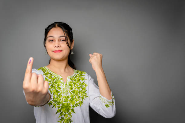 Portrait of happy Indian young woman showing ink-marked finger after voting for the election and looking at the camera with a smile against isolated gray background with copy space.