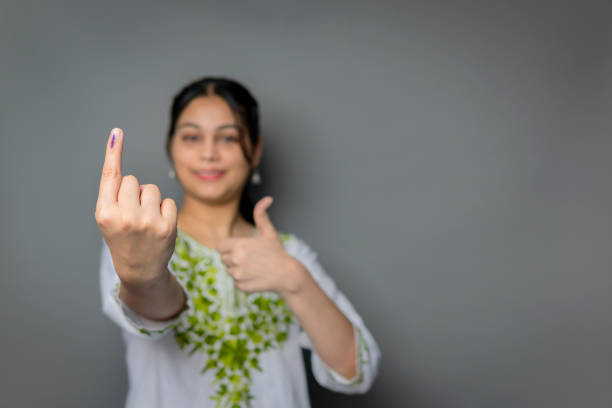 Portrait of happy Indian young woman showing ink-marked finger with thumbs up after voting for the election and looking at the camera with a smile against isolated gray background with copy space.
