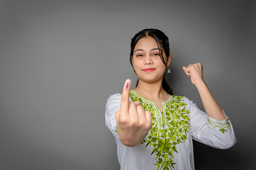 Portrait of happy Indian young woman showing ink-marked finger after voting for the election and looking at the camera with a smile against isolated gray background with copy space.