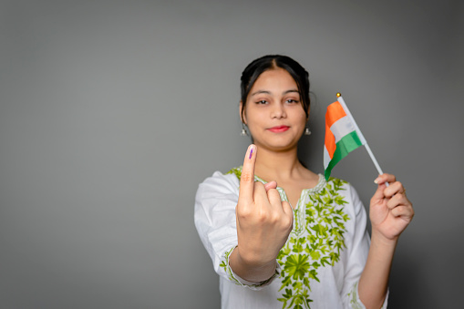 Portrait of happy Indian young woman holding national flag and showing ink-marked finger after voting for the election and looking at the camera with a smile against isolated gray background with copy space.