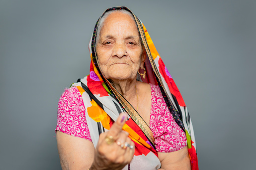 Portrait of happy Indian senior woman in sari showing ink-marked finger after voting for the election and looking at the camera with a smile against isolated gray background with copy space.