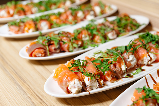 Plates of smoked salmon rolls filled with cream cheese, garnished with arugula and a balsamic glaze, arranged on a wooden table.