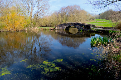 Old stone packhorse bridge over a pond on a canal towpath in the north west of England, UK.