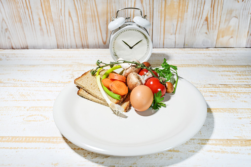 Interval fasting diet concept symbolized by a plate filled to one third with food and an alarm clock on a light wooden background, method for healthy and lasting weight loss, copy space, selected focus