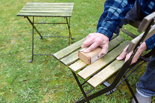 Man is cleaning and restoring wooden outdoor furniture, sanding the weathered wood to remove algae before oiling or painting for a neat garden season, copy space, selected focus