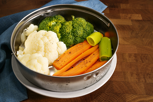 Vegetables such as carrots, broccoli and cauliflower in a metal food steamer basket on a dark rustic wooden table, cooking method to preserve vitamins, minerals and flavor, copy space, selected focus