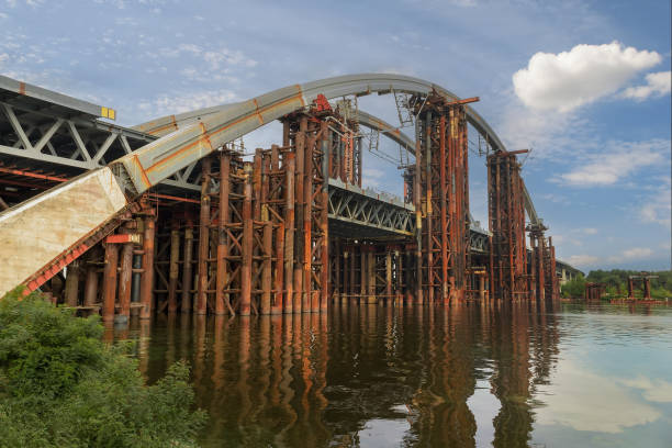 Construction of the bridge across a wide river against a background of a blue sky with white clouds
