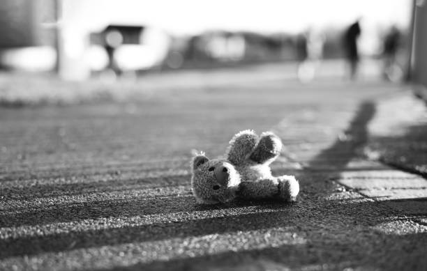 Teddy bear with sad face lying on footpath with blurry people,Image Black,White Lonely bear doll laying down on the road in gloomy day,Lost toy,Loneliness concept, International missing Children