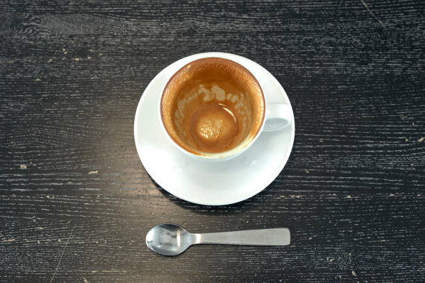 Overhead view of a white cup filled with dark coffee and a spoon on a black textured placemat over a wooden table. Morning light casts shadows, creating a warm and cozy coffee moment.