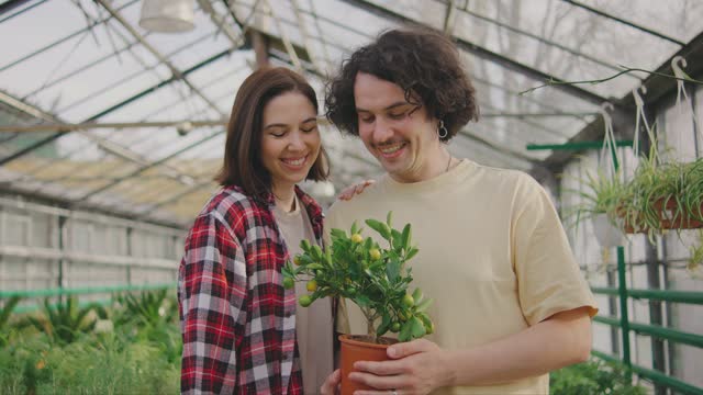 Beautiful young couple in casual clothes holding in their hands a small citrus tree bought in a plant store, looking at camera and smiling