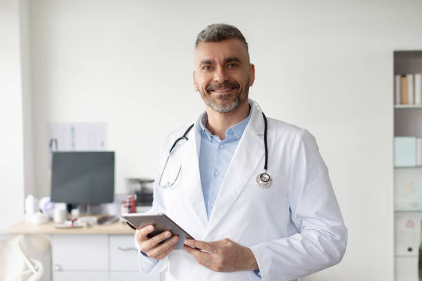 istock Professional help. Happy middle aged male general practitioner standing with tablet and smiling at his office in clinic 2142902602