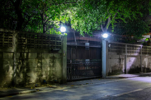 A dark street with a fence and a gate. The gate is lit up with lights. The fence is made of cement blocks
