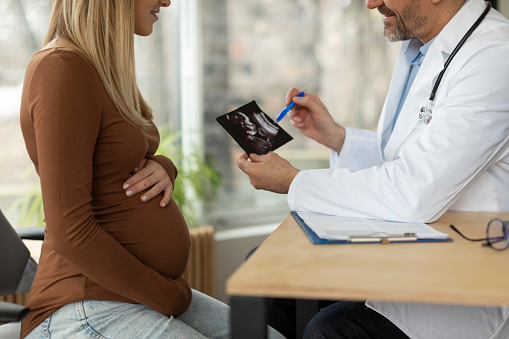 Gynecologist doctor discussing baby sonography image to young pregnant lady during meeting in clinic, doc consulting expectant woman in hospital