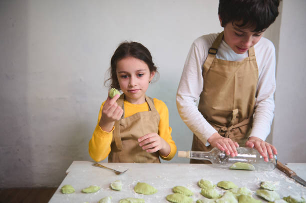 istock Cute little girl stuffing and molding dough while her brother using wine bottle as rolling pin, rolling out a raw green dough. Kids making ravioli, varennyki, pelmeni against white wall background 2142977992