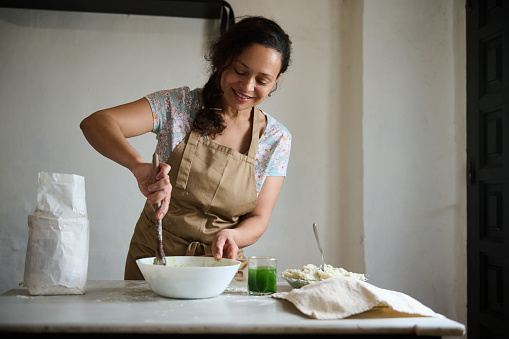 Beautiful authentic woman housewife in beige apron, using wooden spoon, mixing spinach water with whole grain flour for making dough, standing at marble kitchen table in the countryside rural house