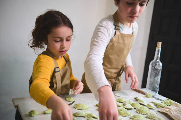 istock Boy and girl making dumplings, standing at floured kitchen table in the rustic kitchen interior. Cooking class for kids. Preparing ravioli, pelmeni, varennyky according to traditional recipe 2143044606