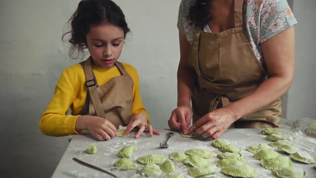 Little girl standing at floured kitchen table, helping her mother to stuff and sculpt dumplings