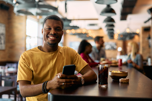 Happy African American man text messaging on smart phone in a pub and looking at camera.