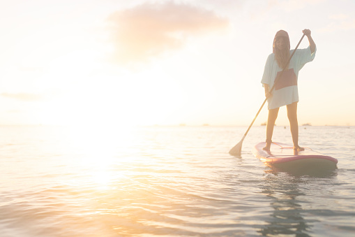 Female tourist standing on stand up paddleboard, paddling, enjoying golden hour