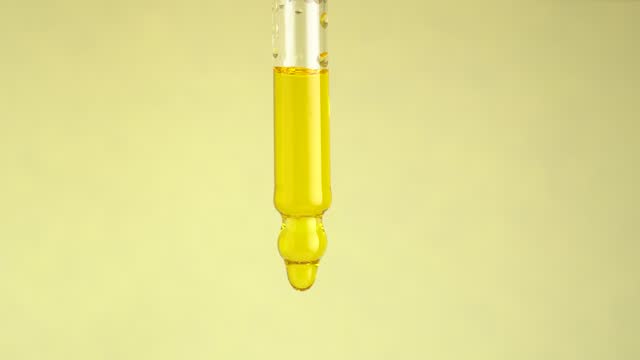 Drops of serum or essential oil fall from a glass pipette on a yellow background, macro shot. Beauty concept.