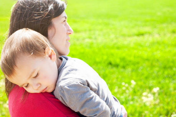 istock Little boy sleeping on his mother's shoulders next to a green spring meadow. 2143431095